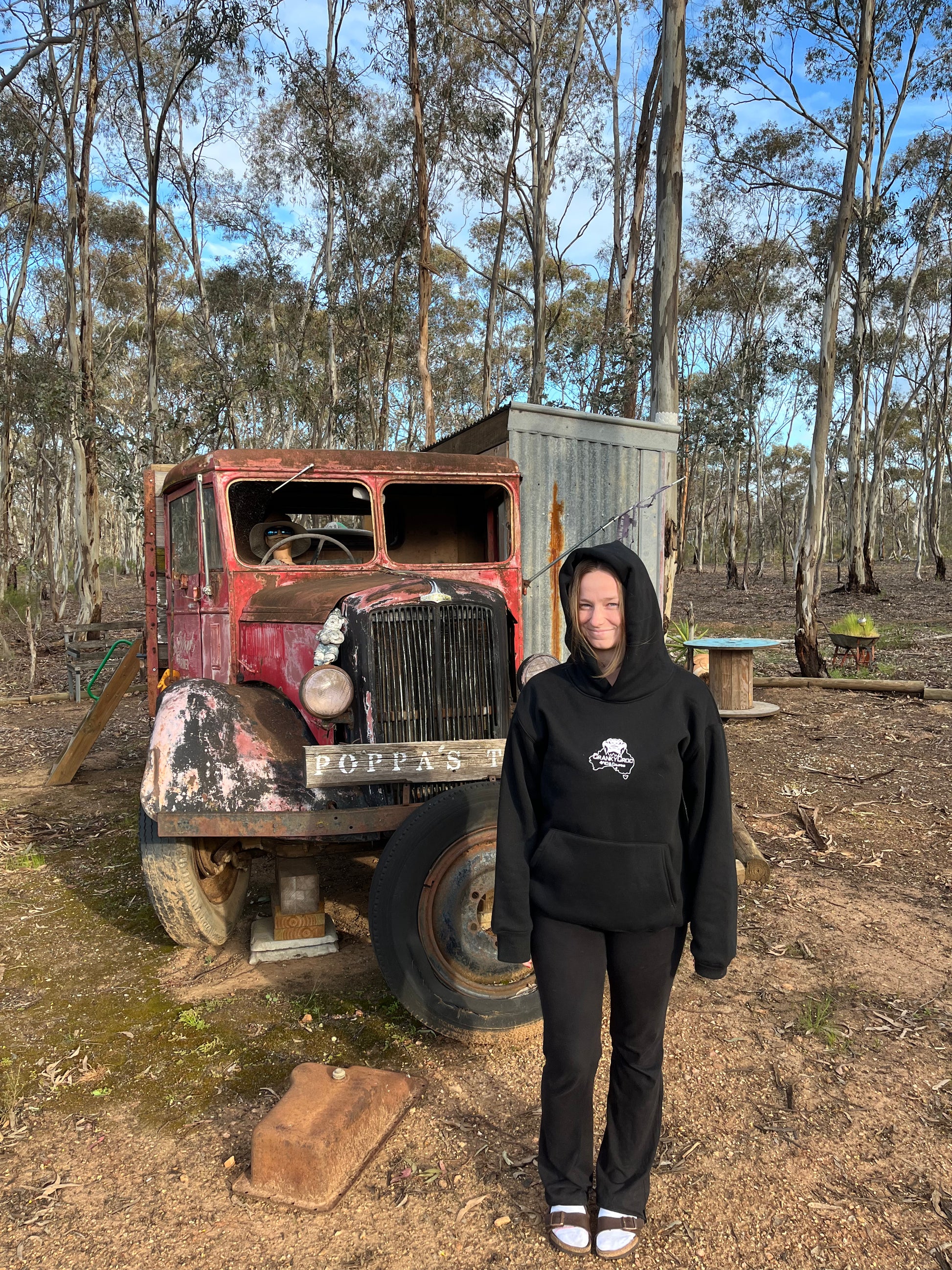 front view of woman wearing a black hoodie standing in front of an old, rusted truck in a wooded area.