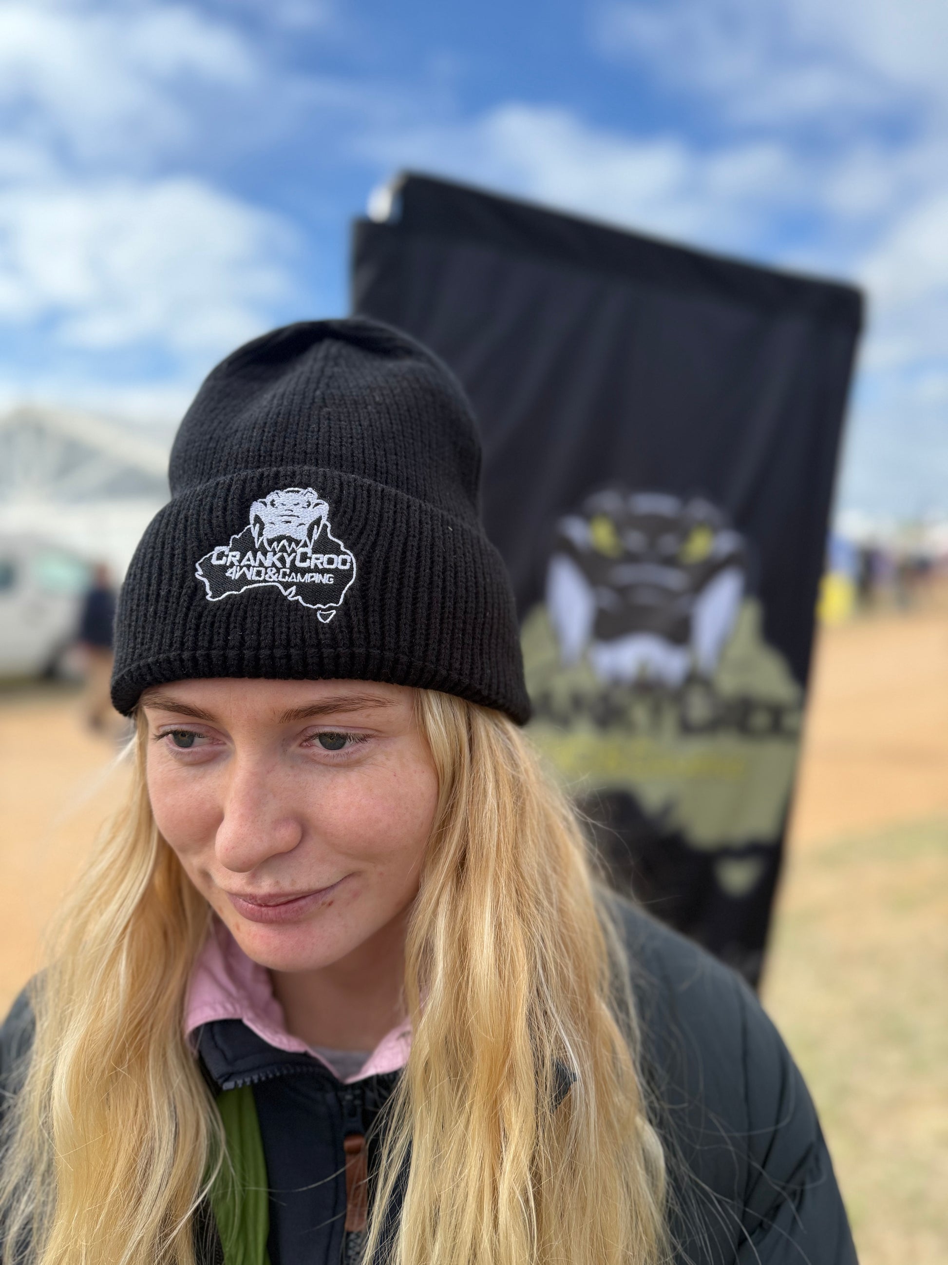 Person wearing a thin black warm beanie with a white embroidered logo, standing outdoors with a branded backdrop.