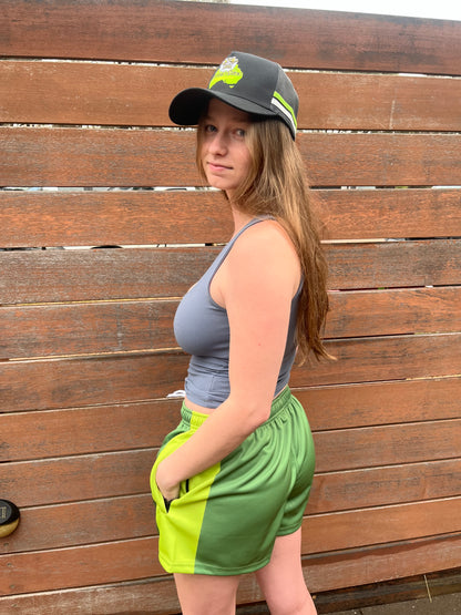 Young girl wearing a black trucker hat with a Cranky Croc logo, The trucker cap also has a green and white stripe on the side. Girl is standing against a wooden fence.