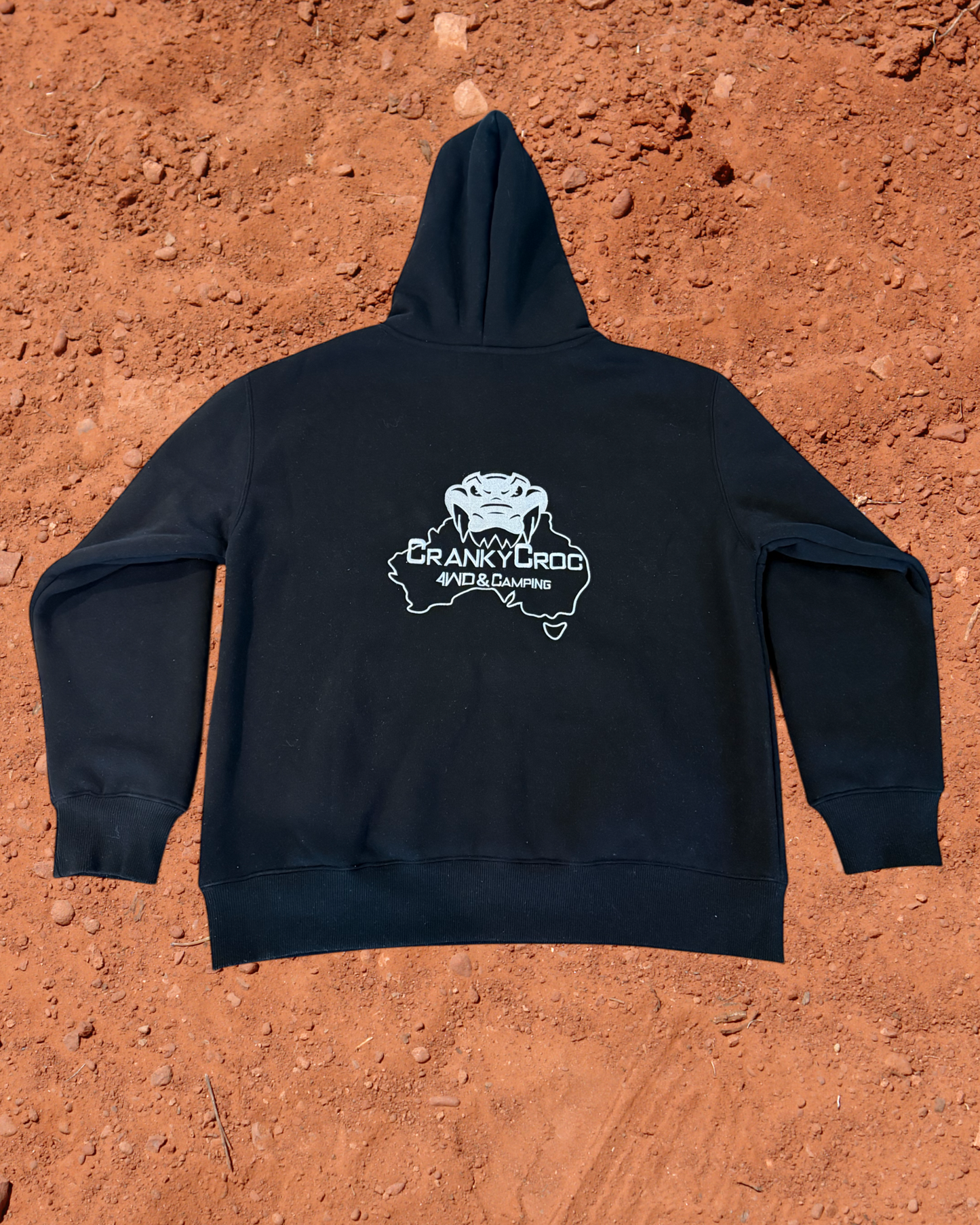 back view of a black hoodie with a white embroidered Cranky Croc logo on the back, placed on a red dirt surface