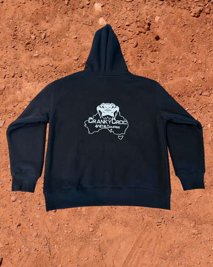 back view of a black hoodie with a white embroidered Cranky Croc logo on the back, placed on a red dirt surface