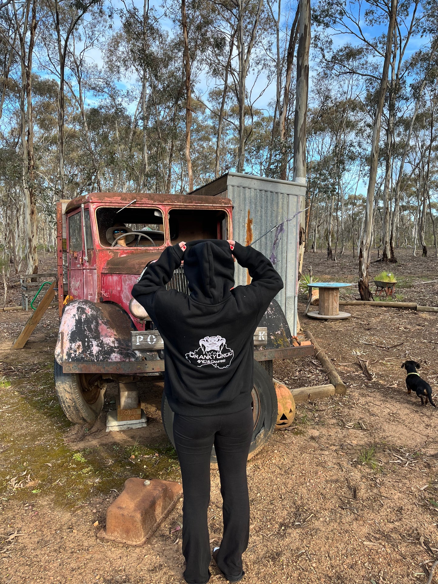 Woman wearing a black hoodie with a logo standing in front of an old truck in a bush land setting.