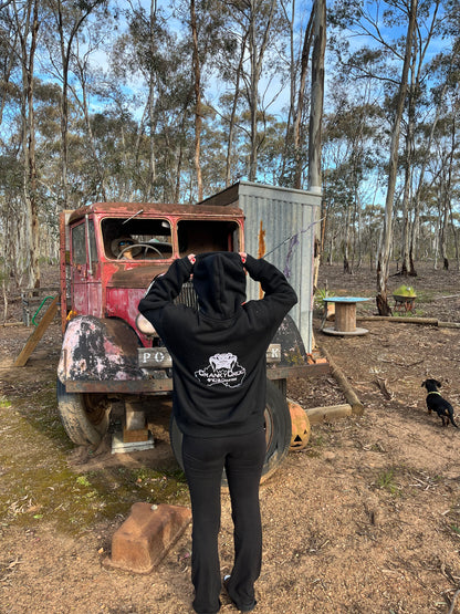 Woman wearing a black hoodie with a logo standing in front of an old truck in a bush land setting.