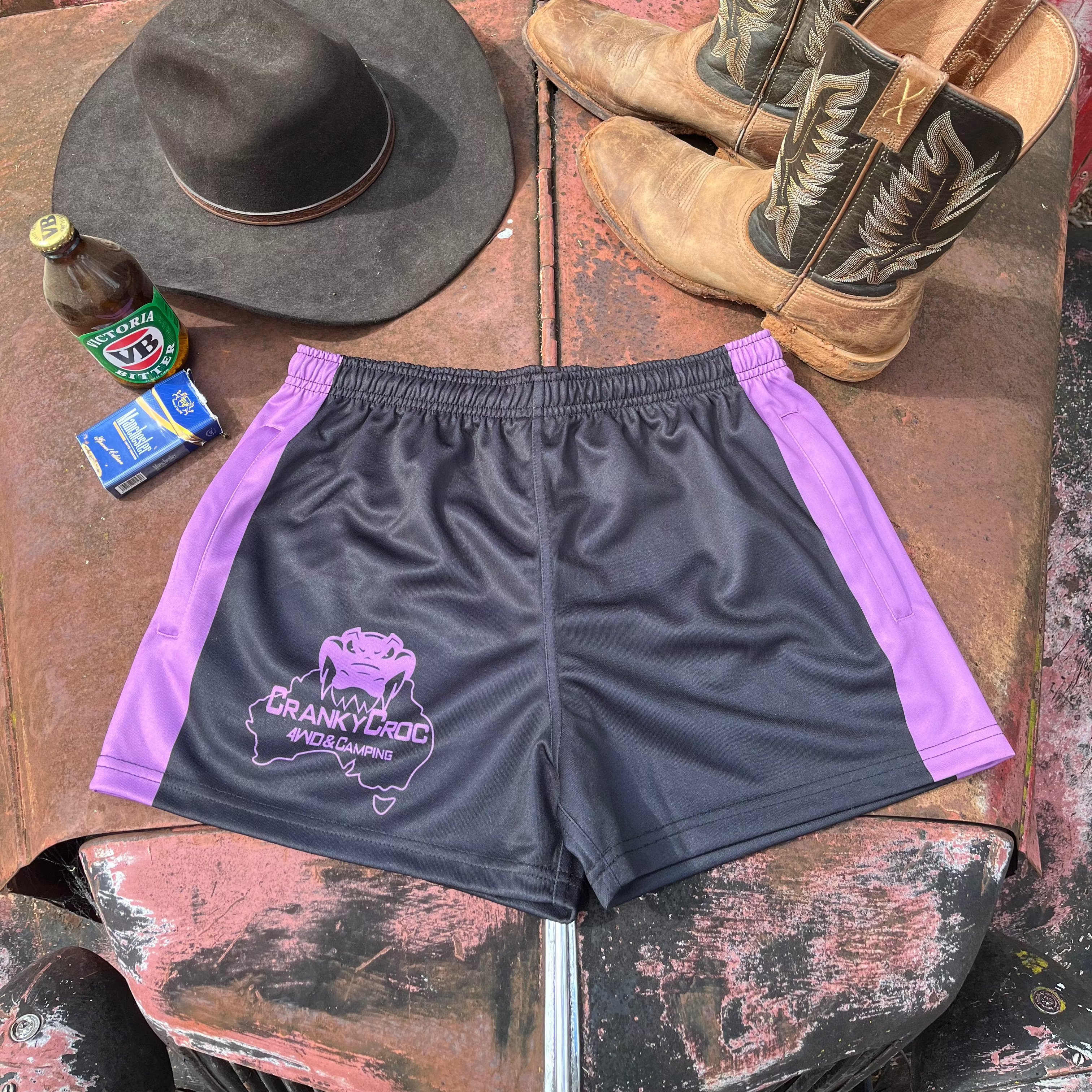 Black with purple Country Aussie Footy Shorts on a 4wd bonnet next to a pair of country boots, country cowboy hat and a beer.