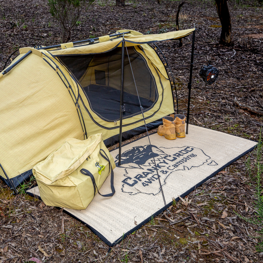 A swag tent set up outdoors on a wooded campsite with a matching a branded ground swag mat rolled out next to it. The tent has a black mesh front window and black support poles, with two tan boots placed near the opening.